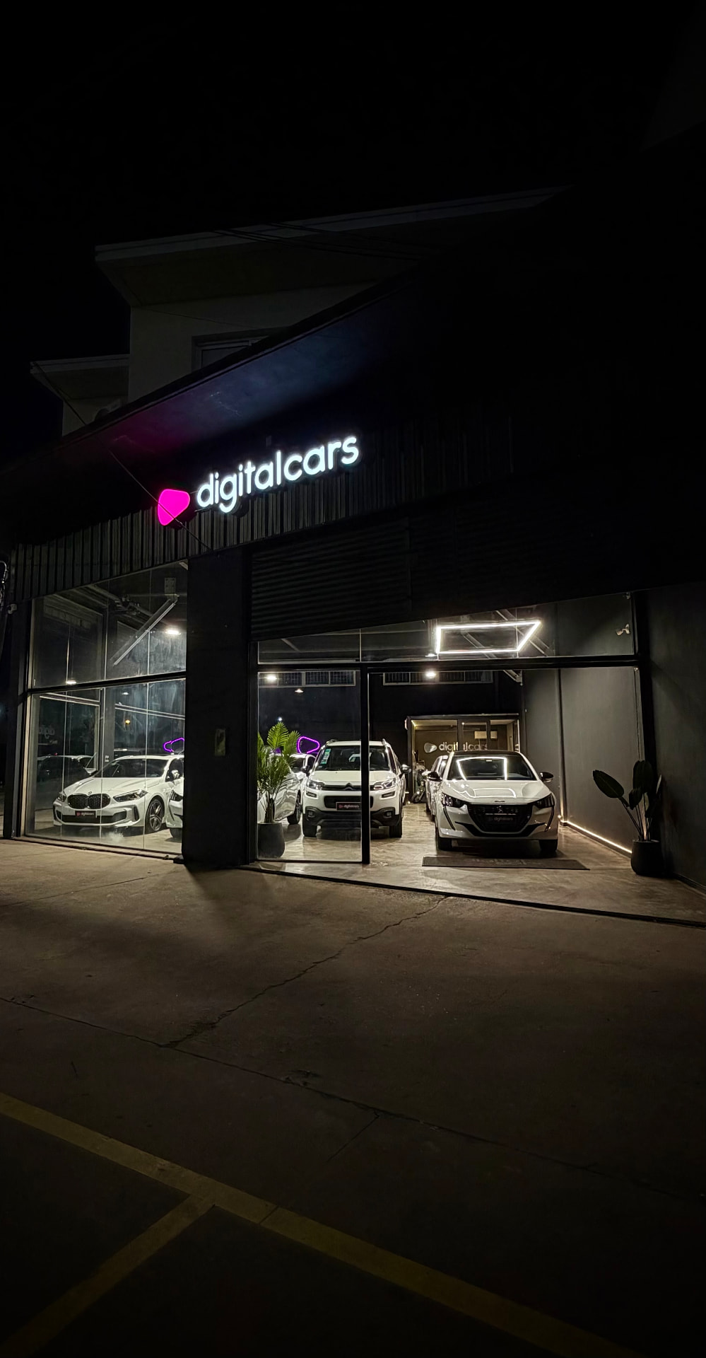 Fachada de la agencia Digital Cars de noche, con autos en exhibición